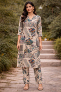 A woman stands outdoors on stone steps wearing a beige printed kurta and matching pants set. The outfit has a patchwork-style pattern with blue, green, and brown motifs on a light beige base.