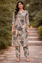 A woman stands outdoors on stone steps wearing a beige printed kurta and matching pants set. The outfit has a patchwork-style pattern with blue, green, and brown motifs on a light beige base.